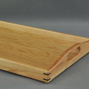May include: A rectangular wooden serving tray with a light brown finish. The tray has cut-out handles on each end and is constructed with visible dovetail joints. The tray is approximately 18 inches long and 12 inches wide.