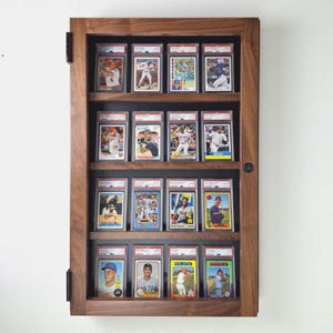 May include: A wooden display case with a dark finish, showcasing multiple baseball cards in protective holders. The cards are arranged in rows, with various players and team logos visible. The case has a hinged door and is mounted on a white wall.