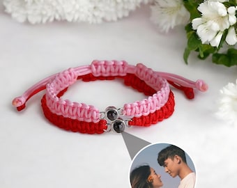 May include: Two red and pink braided friendship bracelets with a silver charm featuring two black stones. The charm is in the shape of two circles.