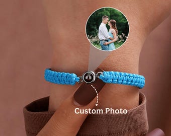 Peut inclure: Un bracelet tressé bleu avec un fermoir magnétique. Le bracelet comporte un petit charme rond noir et argenté. Les mots "Custom Photo" sont écrits en dessous. Une petite photo d'un couple est en arrière-plan.