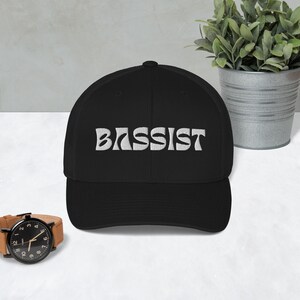 Puede incluir: Una gorra de béisbol negra y blanca con la palabra "BASSIST" bordada en blanco en una fuente retro.