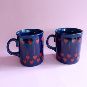 Può includere: Due tazze in ceramica blu con disegni di cuori rossi. Le tazze hanno un interno bianco e un manico arrotondato.