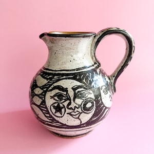 Könnte beinhalten: Ein weißer Keramik Krug mit einem schwarz-weißen Design einer Sonne und eines Mondes mit Gesichtern. Der Krug hat eine gesprenkelte Glasur und einen braunen Rand.