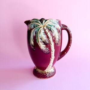 Op de afbeelding: Een bordeauxrode keramische kan met een gedetailleerd palmboomontwerp in wit, groen en goud. De kan heeft een gebogen handvat en een gouden voet. De achtergrond is effen roze.
