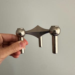 May include: A silver-toned metal candle holder with a geometric design. It features two spherical candle holders connected by a triangular element and a central vertical support. The piece has a modern, minimalist aesthetic.