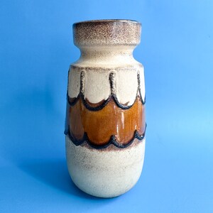 May include: A ceramic vase featuring a cylindrical body with a flared rim. The surface is finished in a cream-colored glaze with a speckled texture. A central band of glossy brown glaze is defined by dark, raised, scalloped outlines.