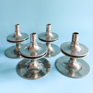 May include: Four silver candle holders. Each has a circular base, a central stem, and a flat disc. The top has a cylindrical opening for a candle. The holders are arranged against a light blue background.