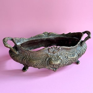 May include: An oval-shaped bronze-toned metal bowl featuring ornate relief detailing, a scalloped rim, and two side handles. The vessel is supported by four small feet and displays a textured, aged finish with intricate patterns across its surface.