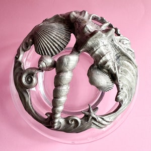May include: A clear glass bowl with a decorative, silver-coloured metal rim featuring seashell and starfish designs. The rim has a spiral design in the centre and a starfish on the bottom. The bowl is set against a pink background.