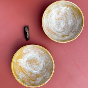 Puede incluir: Dos cuencos de cerámica hechos a mano con borde amarillo e interior blanco arremolinado. Una pequeña pieza de madera oscura se encuentra entre los cuencos. Los cuencos están colocados sobre un fondo rosa polvoriento liso. Los cuencos parecen estar pintados a mano.