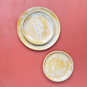 Puede incluir: Dos platos decorativos de madera con un acabado amarillo y blanco envejecido. Los platos son redondos, con un plato más grande y otro más pequeño, sobre un fondo rojo liso. Estilo rústico y campestre.