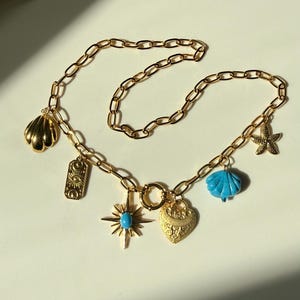May include: Gold-toned chain necklace with various charms, including a seashell, an eye-shaped pendant, a starburst with a turquoise center, a heart, a blue seashell, and a starfish. The necklace is displayed on a white surface.