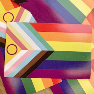 Puede incluir: Una pila de banderas de orgullo coloridas con un círculo amarillo en la esquina de cada bandera. Las banderas están dispuestas de forma que se muestran los diferentes colores y patrones de cada bandera.