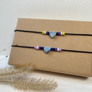 May include: Two beaded bracelets with heart charms. The top bracelet has yellow, white, purple, and black beads. The bottom bracelet has pink, white, purple, and blue beads.