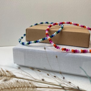 Könnte beinhalten: Zwei bunte Perlenarmbänder auf einer weißen Schachtel. Die Armbänder bestehen aus kleinen, bunten Perlen in verschiedenen Farben, darunter Rosa, Orange, Weiß, Blau und Grün.