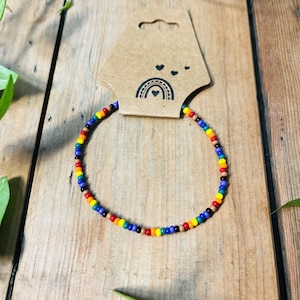 Pode incluir: Um bracelete de contas coloridas do arco-íris com contas de madeira marrom. O bracelete está sobre uma superfície de madeira marrom com uma pequena etiqueta de papel com um arco-íris e corações.