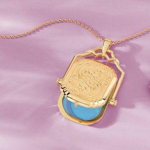 Puede incluir: Collar con dije de oro con un diseño grabado detallado en la parte delantera. El dije está abierto, revelando un interior azul.