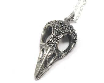 May include: A silver-colored pendant necklace featuring a detailed raven skull design. The skull has intricate floral patterns on the top and sides, with a long, pointed beak. The necklace chain is delicate and silver.