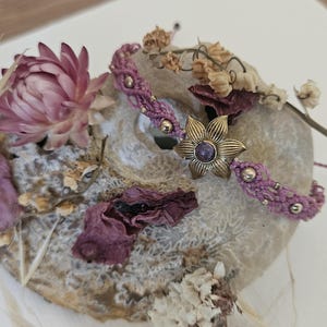 May include: A handcrafted macrame bracelet in shades of purple, featuring a brass flower-shaped centerpiece with a purple stone. The bracelet is adorned with small gold beads. The image also includes dried flowers and petals.