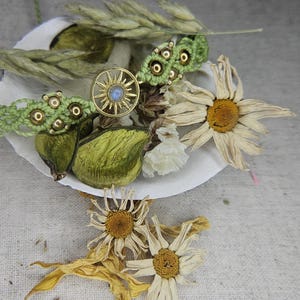 Peut inclure: Une bague dorée avec un motif de soleil et une pierre bleue au centre, accompagnée d'un bracelet macramé vert, de fleurs séchées et de gousses, disposés sur une coquille blanche. Le fond est en tissu de lin neutre.
