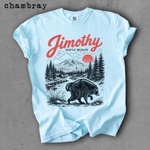 May include: A light blue t-shirt featuring a graphic print with the red text "Jimothy" above "SEATTLE WILDLIFE". The illustration depicts a black raccoon in the foreground, a forest of pine trees, a mountain range, and a red sun in the sky.