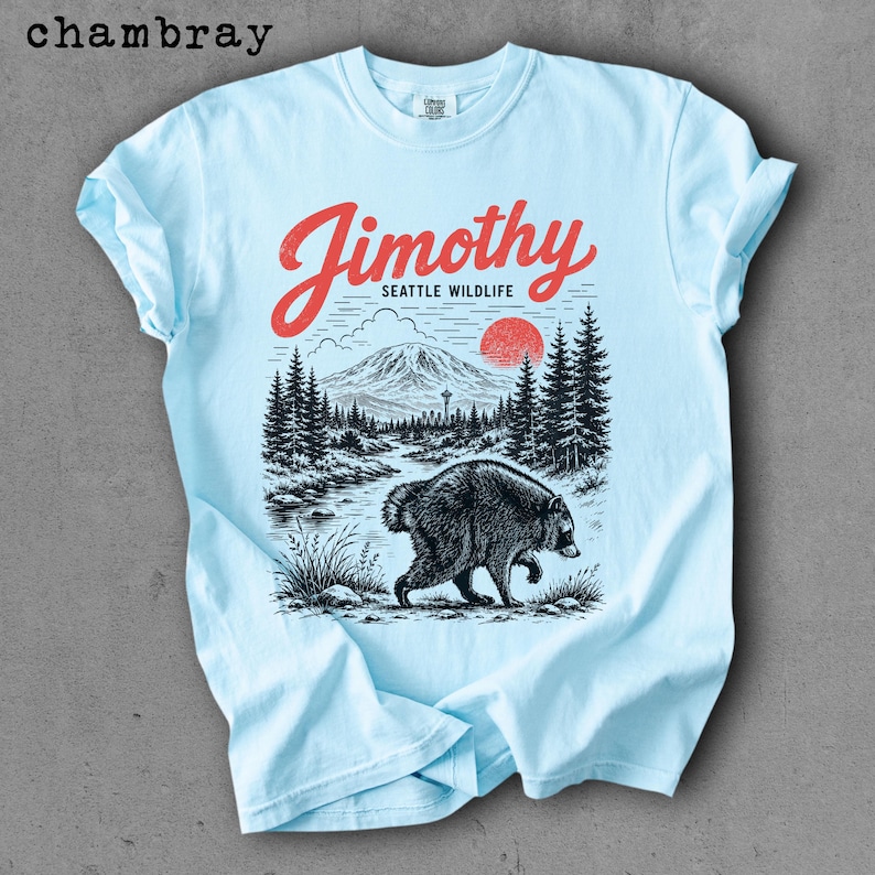 May include: A light blue t-shirt featuring a graphic print with the red text "Jimothy" above "SEATTLE WILDLIFE". The illustration depicts a black raccoon in the foreground, a forest of pine trees, a mountain range, and a red sun in the sky.
