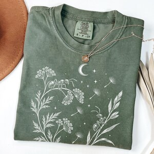 Op de afbeelding: Een olijfgroen t-shirt met een witte grafische print van twee mieren op wilde bloemen, een maansikkel en verspreide sterren. Het shirt heeft een ronde hals en een Comfort Colors-label aan de binnenkant van de kraag met de maat small.