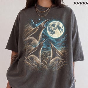 Könnte beinhalten: Ein anthrazitfarbenes T-Shirt mit einem Grafikdruck von drei Ameisenbären, die einen großen, hellen Vollmond in einem sternenklaren Nachthimmel über einem Feld aus hohem Gras anheulen. Das Wort PEPPER steht in der oberen rechten Ecke des Bildes.