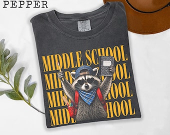 Może przedstawiać: Ciemnoszary t-shirt z grafiką szopa pracza w niebieskiej czapce z daszkiem, okularach przeciwsłonecznych w kształcie serc i niebieskiej bandanie w kratę. Szop ma czerwony plecak, ołówek i notatnik. Napis MIDDLE SCHOOL jest wydrukowany w kolorze żółtym.