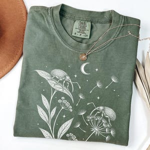 以下が含まれることがあります： オリーブグリーンのTシャツ。白い線画で描かれた2匹のゾウムシが植物の茎にとまり、その周りにタンポポの種、星、三日月が浮かんでいるデザイン。クルーネックで、Comfort Colorsのブランドラベルが付いている。