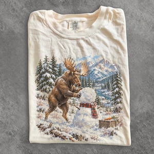 Może przedstawiać: Kremowy t-shirt z krótkim rękawem z nadrukiem przedstawiającym brązowego łosia budującego bałwana w zaśnieżonym górskim krajobrazie z sosnami i rzeką. Koszulka posiada metkę marki Comfort Colors nadrukowaną po wewnętrznej stronie przy szyi.