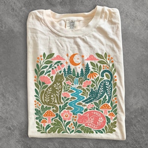 Może przedstawiać: Kremowy t-shirt z nadrukiem graficznym przedstawiającym trzy koty w lesie z wodospadem, półksiężycem, kwiatami i grzybami. Wzór wykorzystuje paletę zieleni, różu, pomarańczu i błękitu. Wewnętrzna metka przy kołnierzu zawiera napis Comfort Colors.