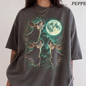 Peut inclure: Un t-shirt gris anthracite à manches courtes avec une impression graphique représentant trois okapis hurlant à la pleine lune dans une forêt sombre. Le mot PEPPER est imprimé dans le coin supérieur droit. Le t-shirt a une coupe décontractée.