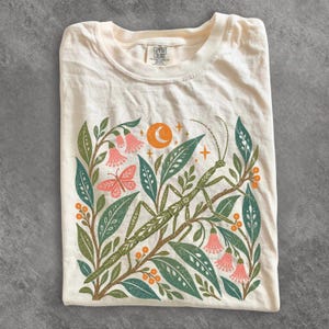 May include: A cream-coloured t-shirt featuring a graphic print of a green praying mantis surrounded by pink bell-shaped flowers, green leaves, orange berries, a butterfly, and a crescent moon with stars. The inner neck label reads Comfort Colors.
