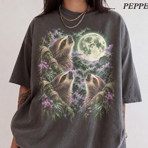 May include: A charcoal grey t-shirt featuring a graphic print of three sloths looking up towards a full moon in a forest setting with purple flowers and green foliage. The word PEPPER is printed in the upper right corner of the garment.