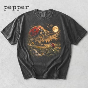 Può includere: Una t-shirt grigio antracite a maniche corte con una stampa grafica di due coccinelle rosse in un paesaggio boschivo al chiaro di luna con un ruscello e fiori. La parola pepper è stampata in nero nell'angolo in alto a sinistra della maglia.