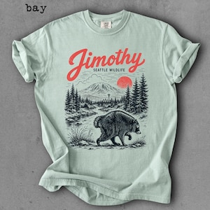 May include: A light green t-shirt featuring a graphic print of a bear walking through a forest landscape with a mountain, a red sun, and the text Jimothy Seattle Wildlife in red and black ink. The shirt has a crew neckline and short sleeves.