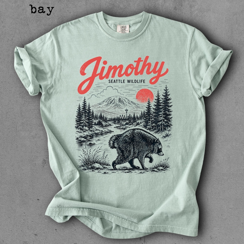 May include: A light green t-shirt featuring a graphic print of a bear walking through a forest landscape with a mountain, a red sun, and the text Jimothy Seattle Wildlife in red and black ink. The shirt has a crew neckline and short sleeves.