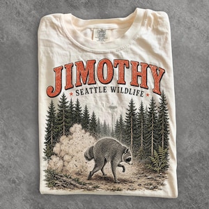 Puede incluir: Una camiseta de color crema con el texto naranja JIMOTHY SEATTLE WILDLIFE sobre un gráfico de un mapache en un bosque con la Space Needle al fondo. La camiseta tiene cuello redondo y una etiqueta de la marca Comfort Colors en el cuello.