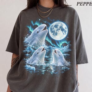 Puede incluir: Una camiseta gris carbón con un estampado gráfico de tres ballenas beluga blancas emergiendo de aguas heladas bajo una luna llena y auroras boreales verdes. La palabra PEPPER está impresa en la esquina superior derecha de la prenda.