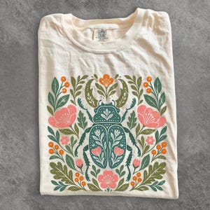 Op de afbeelding: Een crèmekleurig T-shirt met korte mouwen en een centrale afbeelding van een blauwgroene vliegend hert, omringd door symmetrische bloem- en plantmotieven in roze, oranje en groen. Het label in de nek bevat de tekst Comfort Colors.