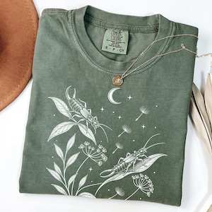 Może przedstawiać: Oliwkowozielony t-shirt z białą grafiką liniową przedstawiającą dwa skorki na liściach, półksiężyc, gwiazdy i nasiona mniszka lekarskiego. Koszulka ma okrągły dekolt i nadrukowaną metkę marki Comfort Colors po wewnętrznej stronie kołnierza.