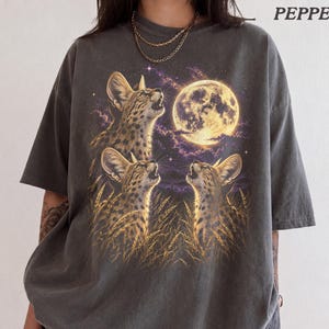 Puede incluir: Una camiseta gris carbón con un estampado gráfico de tres servales en hierba alta mirando hacia una luna llena grande y brillante en un cielo estrellado de color púrpura. La palabra PEPPER está impresa en la esquina superior derecha.
