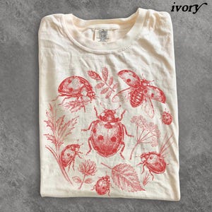 May include: An ivory t-shirt featuring a red graphic print of multiple ladybugs in various poses, interspersed with detailed botanical illustrations of leaves and small flowers. The shirt has a crew neckline and a Comfort Colors brand label.