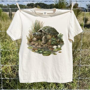 May include: A cream-coloured short-sleeve t-shirt featuring a detailed graphic print of a snapping turtle resting on a small grassy island surrounded by water, reeds, and lily pads. The shirt has a crew neckline and a standard fit.