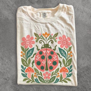 Può includere: Una t-shirt color crema con una grafica centrale raffigurante una coccinella circondata da fiori rosa, foglie verdi, bacche arancioni e due piccoli funghi. L'etichetta del marchio Comfort Colors è visibile all'interno del colletto.