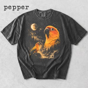 May include: A charcoal gray t-shirt featuring the word pepper in the top left corner. The graphic depicts two orange and yellow parrots in a forest landscape at night under a full moon, with a starry sky and a small waterfall in the background.
