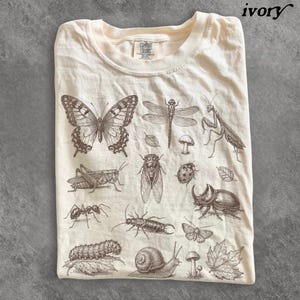 May include: An ivory t-shirt featuring a brown ink illustration of various insects and nature elements, including a butterfly, dragonfly, praying mantis, grasshopper, cicada, ladybug, beetle, ant, caterpillar, snail, mushrooms, and leaves.