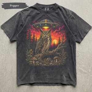 Może przedstawiać: Ciemnoszary t-shirt z nadrukiem graficznym przedstawiającym sowę siedzącą na gałęzi drzewa pod świecącym UFO. Scena zawiera zachód słońca, sylwetki sosen i grzyby. Słowo Pepper jest nadrukowane w lewym górnym rogu.