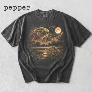 May include: A pepper-coloured short-sleeve t-shirt featuring a graphic print of a large insect with iridescent eyes looming over a swampy landscape under a full moon. The scene includes trees, tall grasses, and a smaller insect resting on a plant.